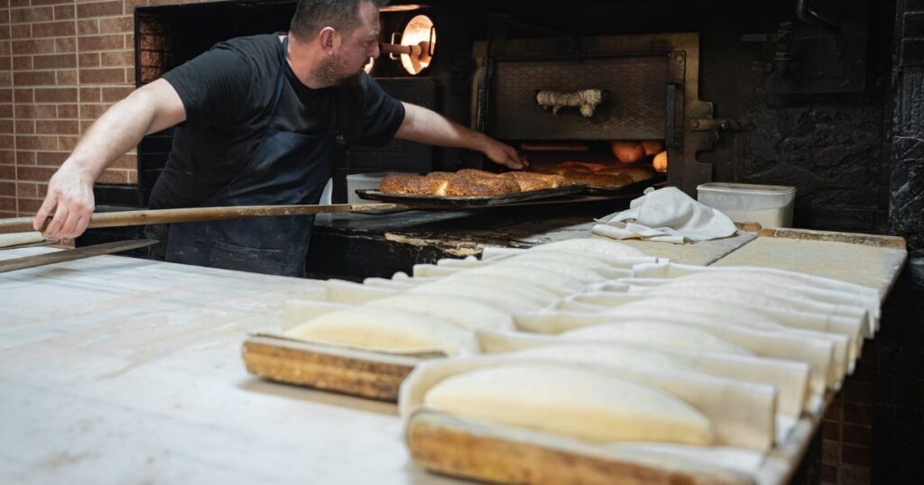 A baker placing fresh bread in a traditional wood-fired oven in a small bakery.