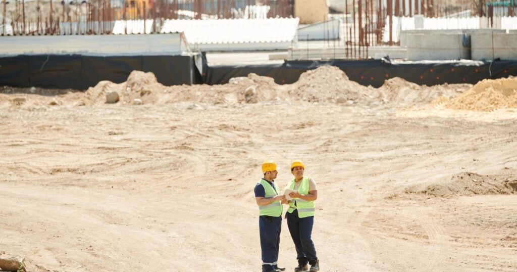 Two construction workers in safety gear discussing plans on a job site.